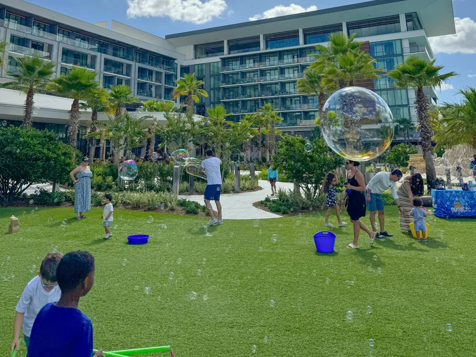 Kids and parents chase giant bubbles on a resort-style lawn beneath palm trees