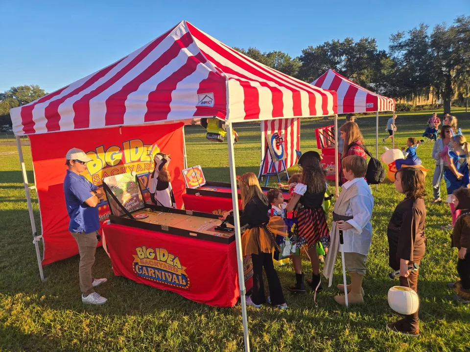 Kids in costumes and parents gather at carnival game tables under striped tents on a sunny lawn