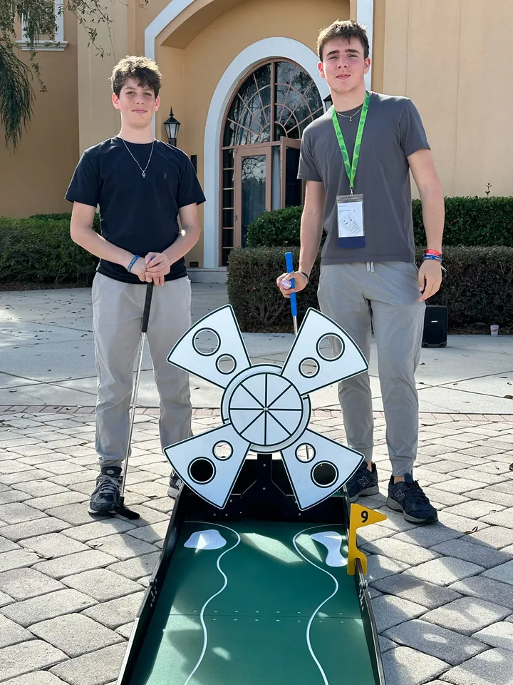Two young adults with event lanyards stand with putters beside a portable mini golf hole