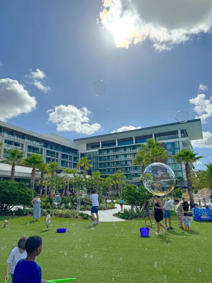 Kids and parents chase giant bubbles on a resort-style lawn beneath palm trees